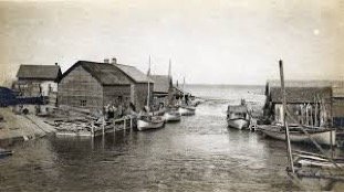 Original Mackinaw boats are moored on the south (left) side the river. The first generation of enclosed fish tugs are tied up on the north side.